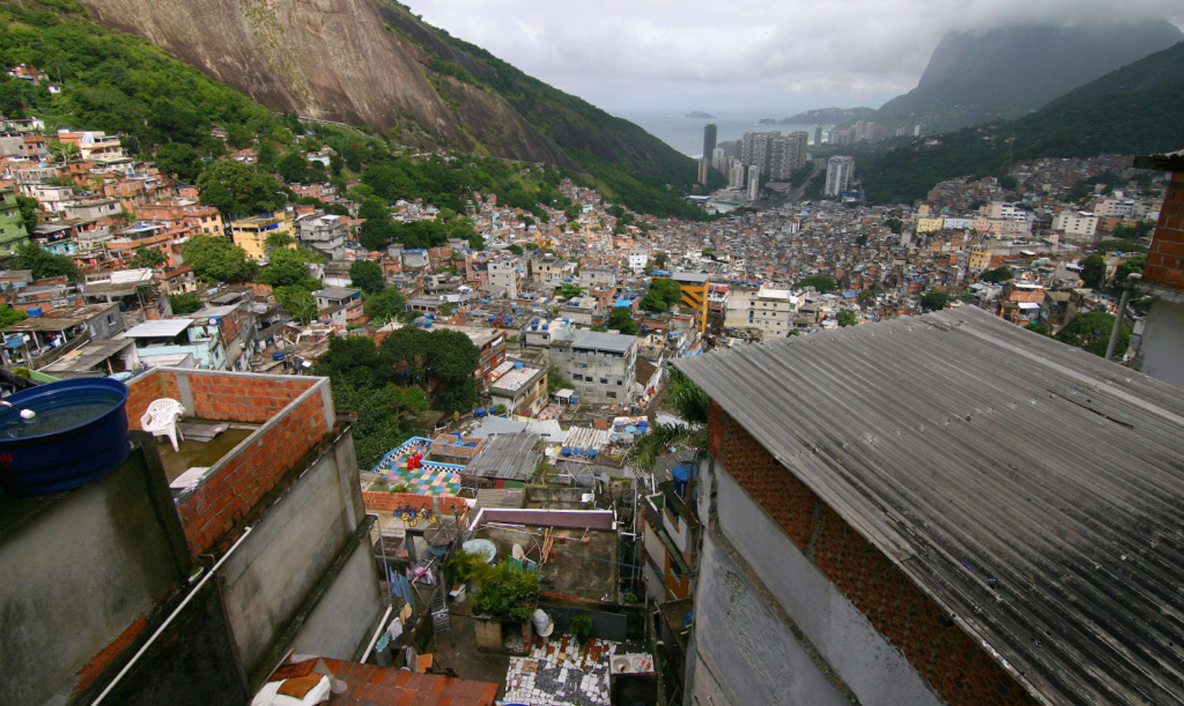 A panoramic view of a densely populated hillside community with colorful buildings, overlooking a coastal area and mountains in the background. The sky is cloudy, suggesting an overcast day.