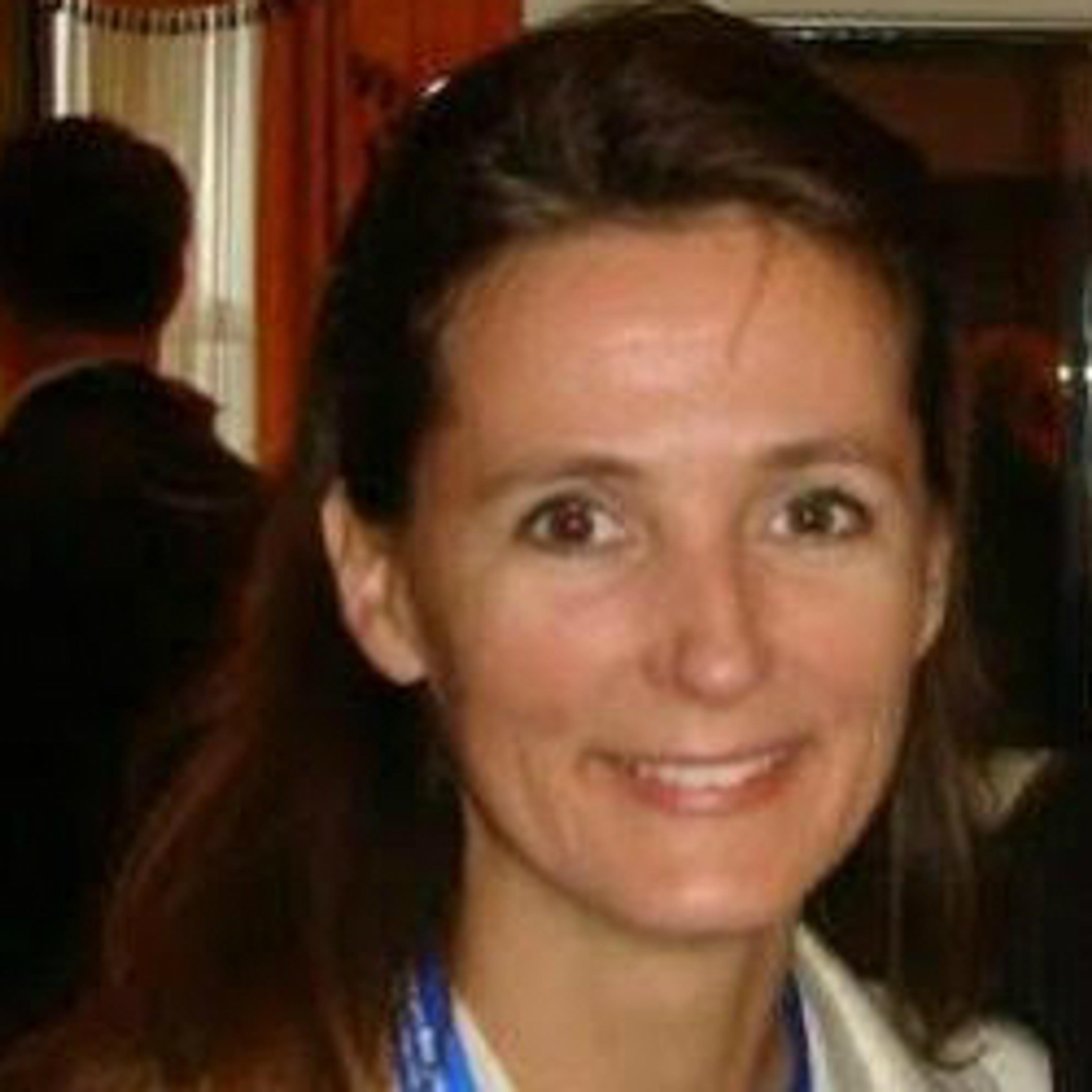 A woman with long brown hair smiles at the camera, wearing a blue lanyard around her neck. The background is blurred with people and indoor decor.