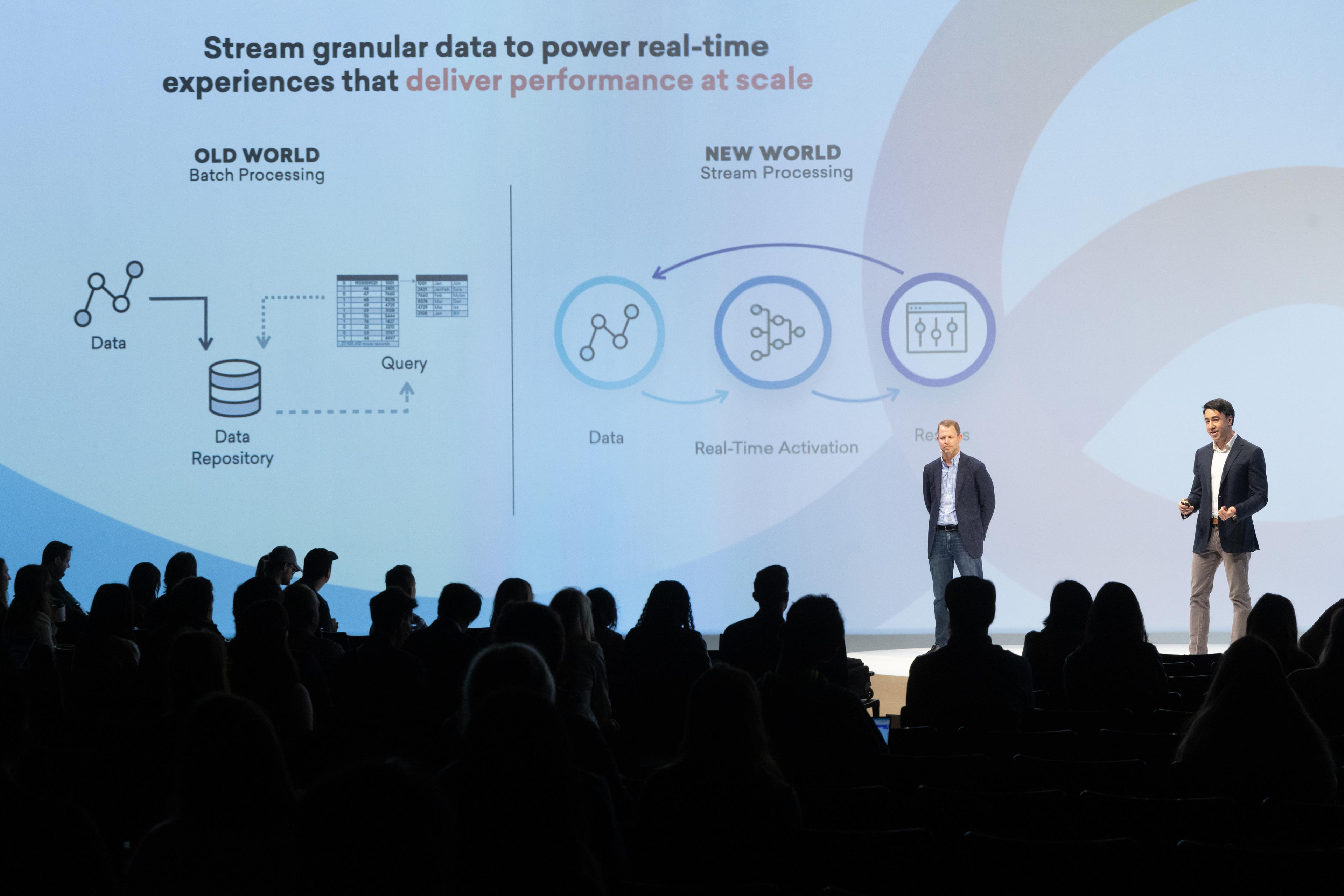 Two presenters stand on stage in front of an audience, discussing the transition from batch processing to real-time data streaming, illustrated by diagrams on a screen.