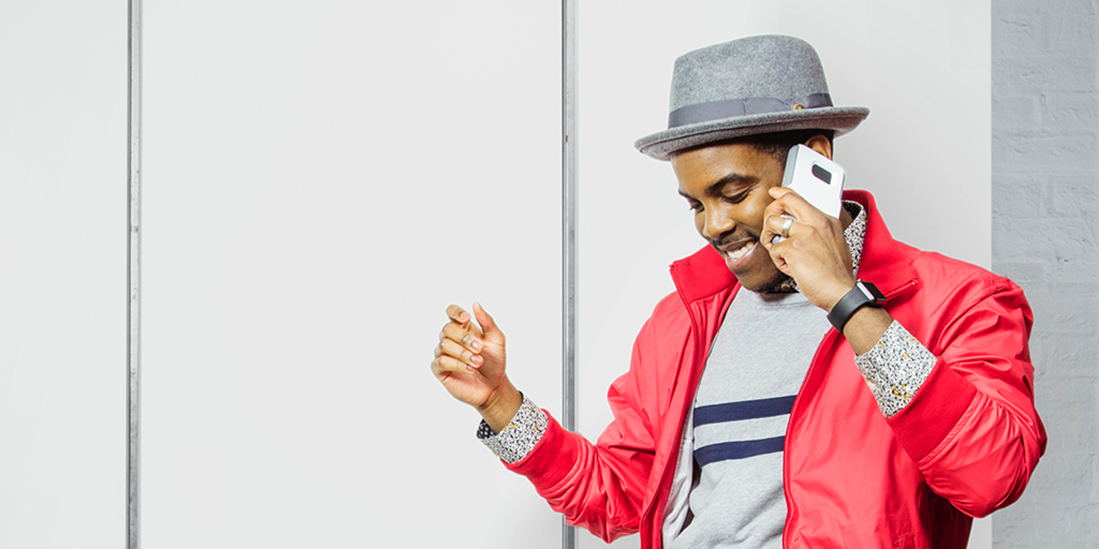 A man in a red jacket and gray hat is smiling while talking on a smartphone. He appears to be enjoying a conversation.