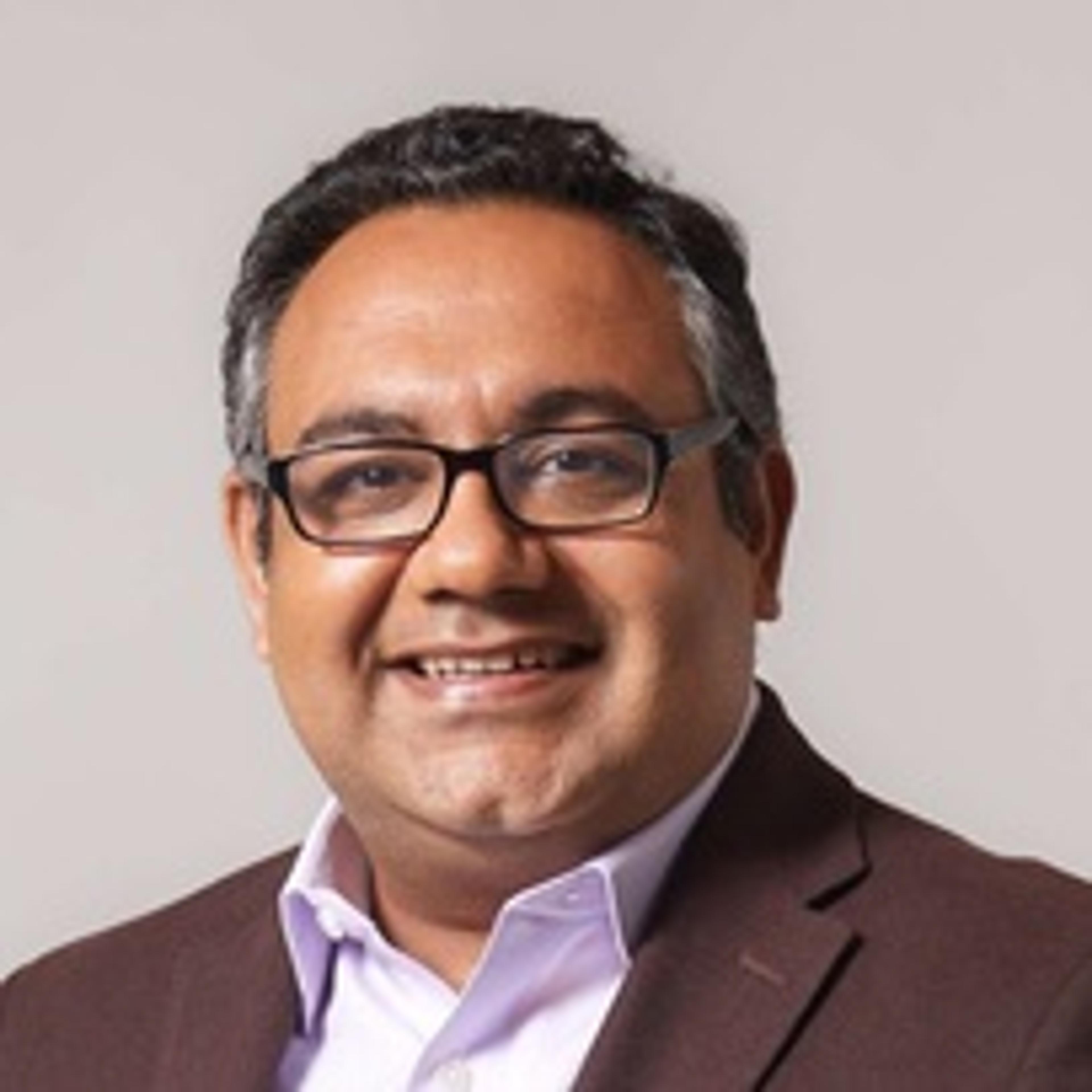 A smiling man with glasses and short dark hair, wearing a dark blazer over a light shirt, poses against a neutral background.