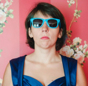 A woman wearing a blue dress and sunglasses stands against a floral wallpaper background, looking directly at the camera. Her expression is serious.