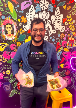 Smiling man holds two plates of tacos in front of a colorful floral mural.