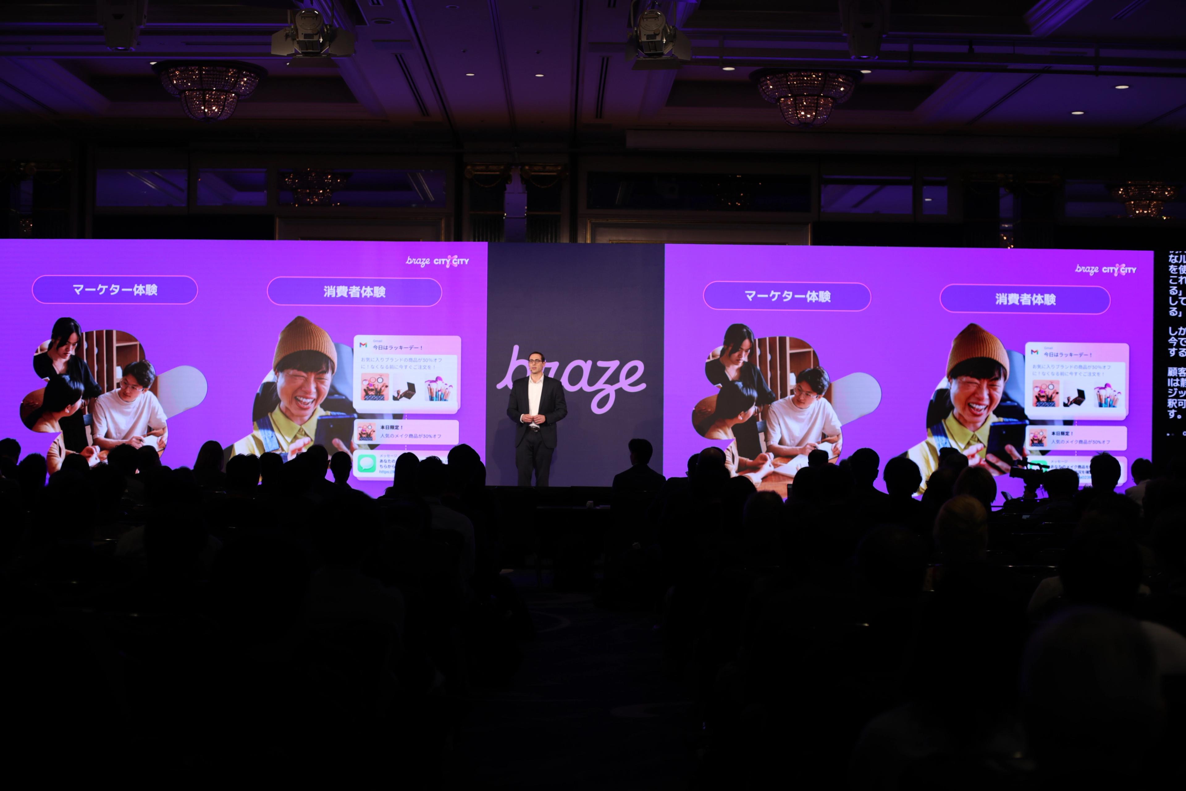 ステージ上で、ブラズのロゴ、デバイスを使用する人々の紫色のグラフィック、日本語テキストを表示する大型スクリーンの横に立つプレゼンター(ジョンハイマン)が、観客に向かって話している。