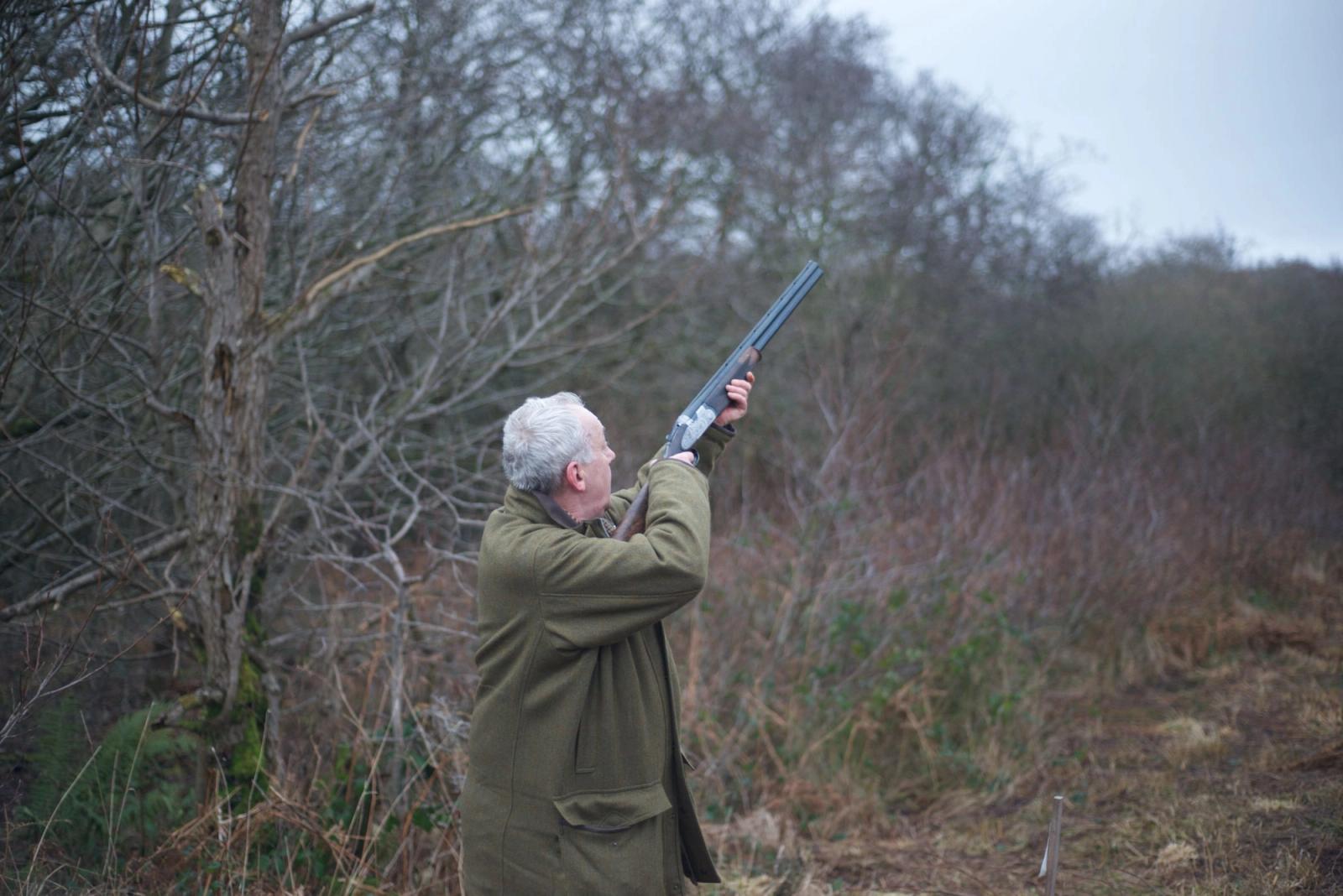 A shooter in a tweed coat raising a shotgun in winter woodland.