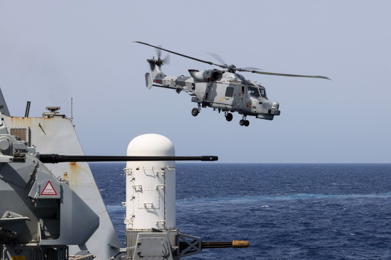 A Royal Navy Wildcat helicopter hovering alongside a warship at sea.