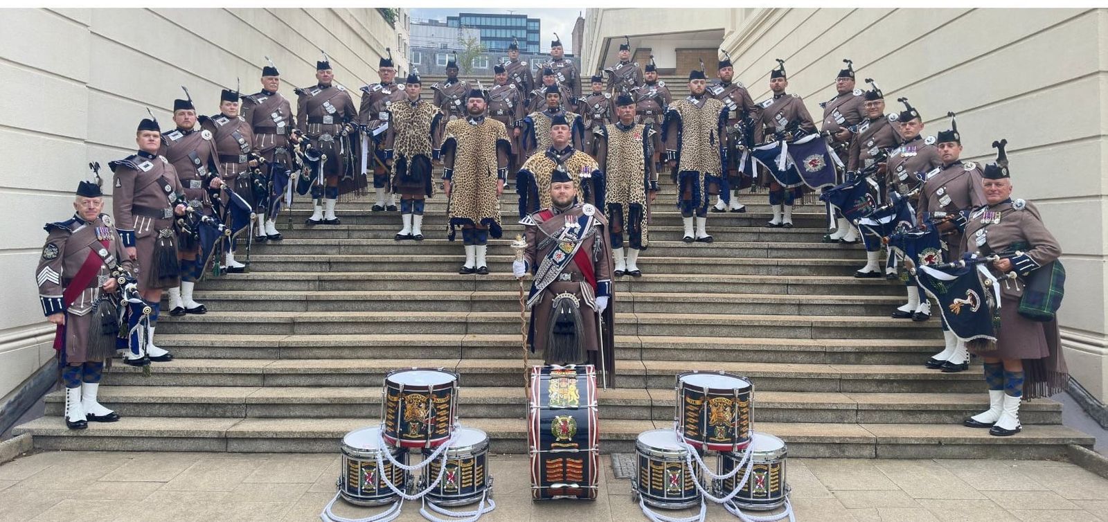 The London Scottish Pipes and Drums in Hodden Grey uniform on ceremonial steps, with drums in the foreground.