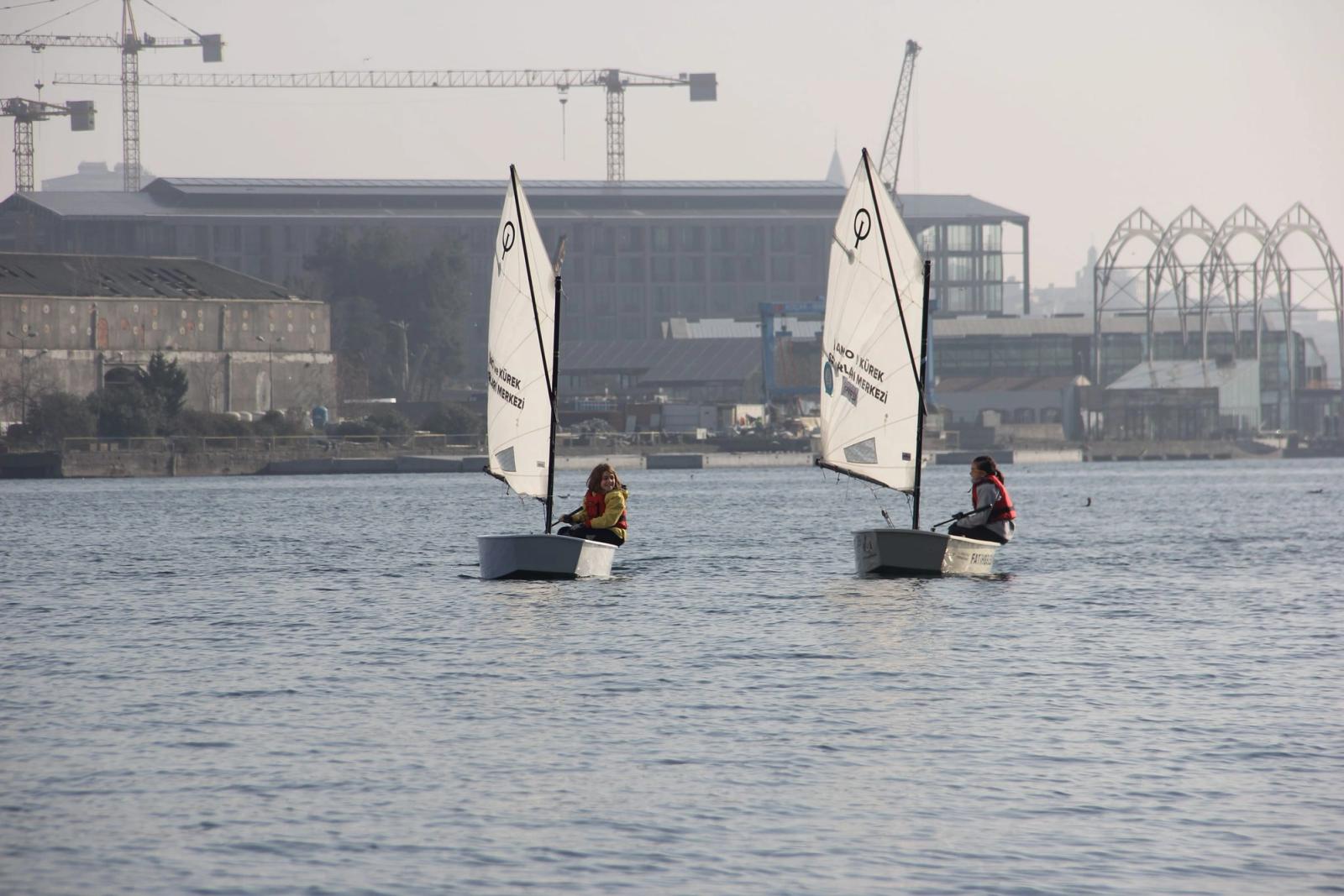 Two sailors in dinghies on calm water, with a harbour and city buildings behind them.