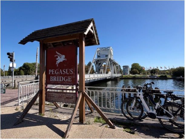 The Pegasus Bridge sign beside the canal, with the bridge and bicycles beyond
