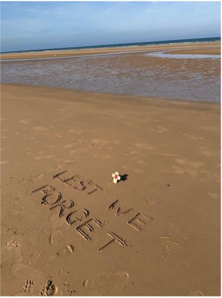 The words Lest we forget written in the sand at Omaha Beach, with a small cross and poppy