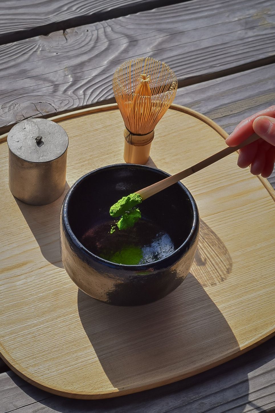 Person stirring green tea in a black bowl with a wooden whisk on a wooden tray.