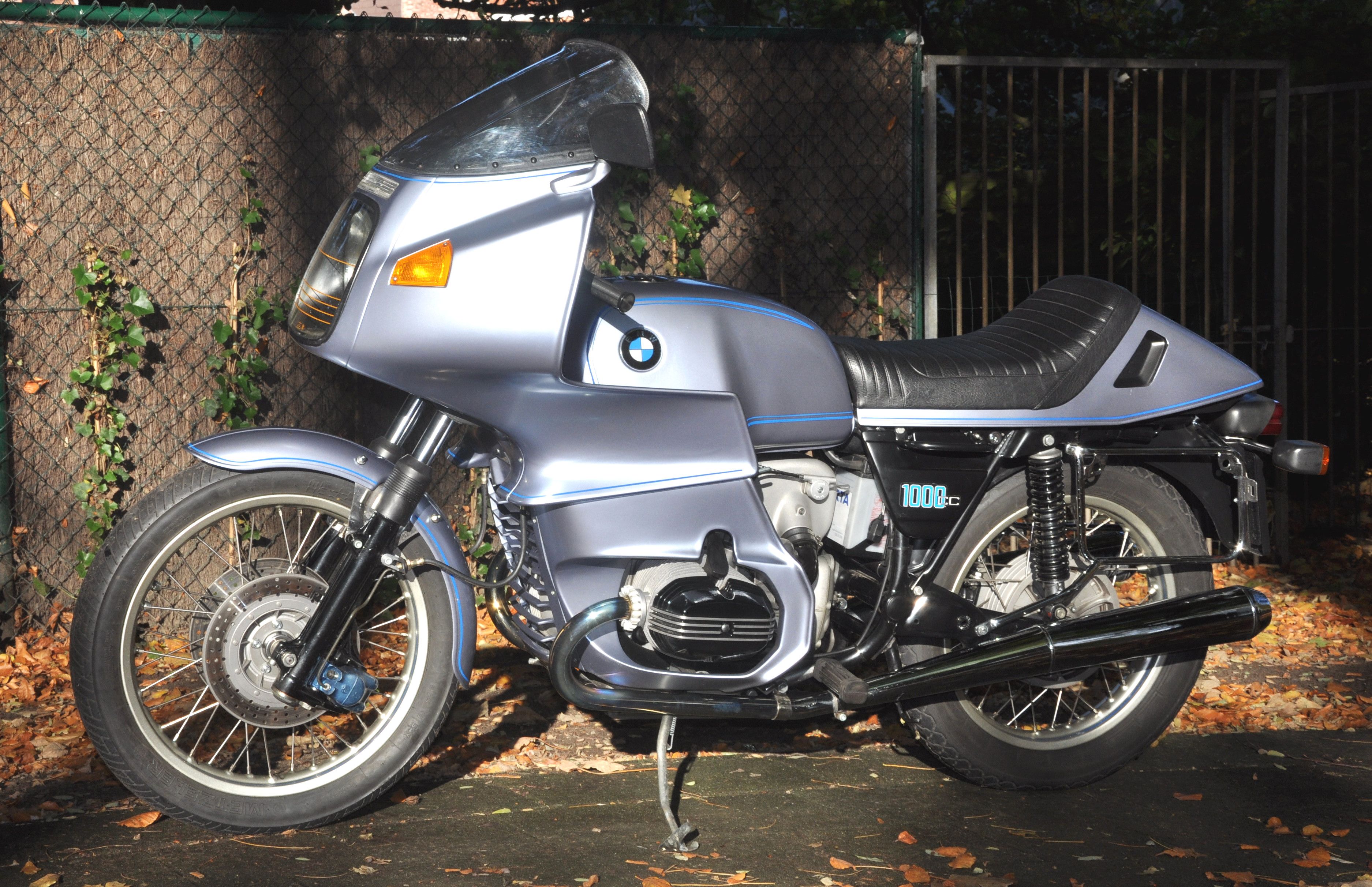 Silver-blue 1977 BMW R100RS with full fairing, side view, parked in front of a stone wall
