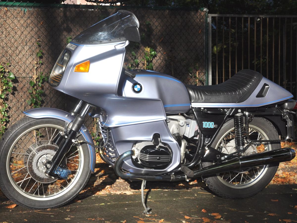 Silver-blue 1977 BMW R100RS with full fairing, side view, parked in front of a stone wall
