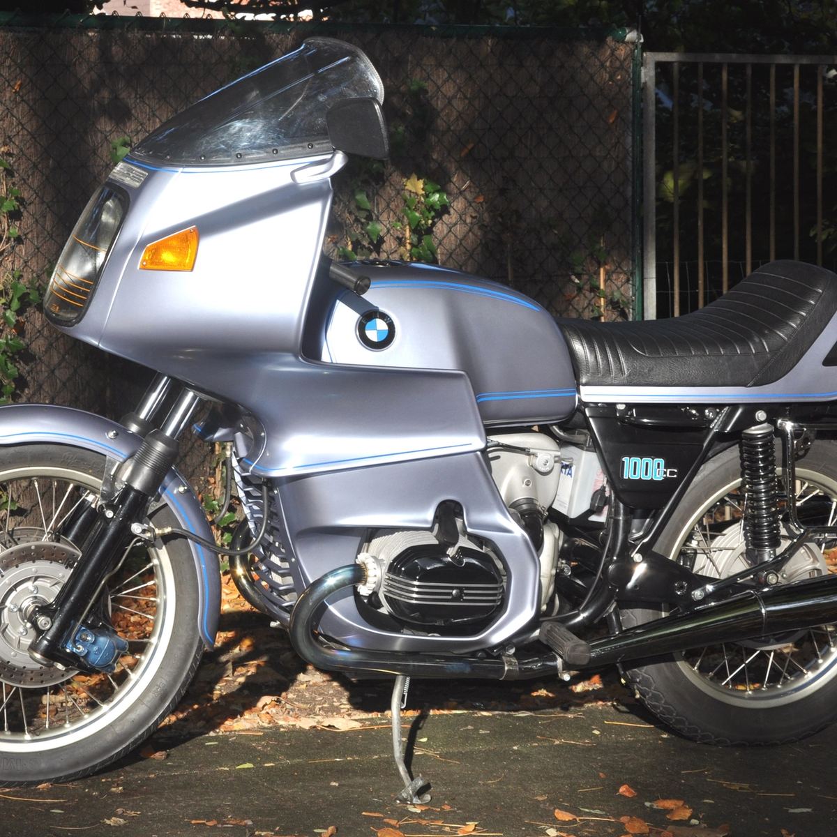 Silver-blue 1977 BMW R100RS with full fairing, side view, parked in front of a stone wall