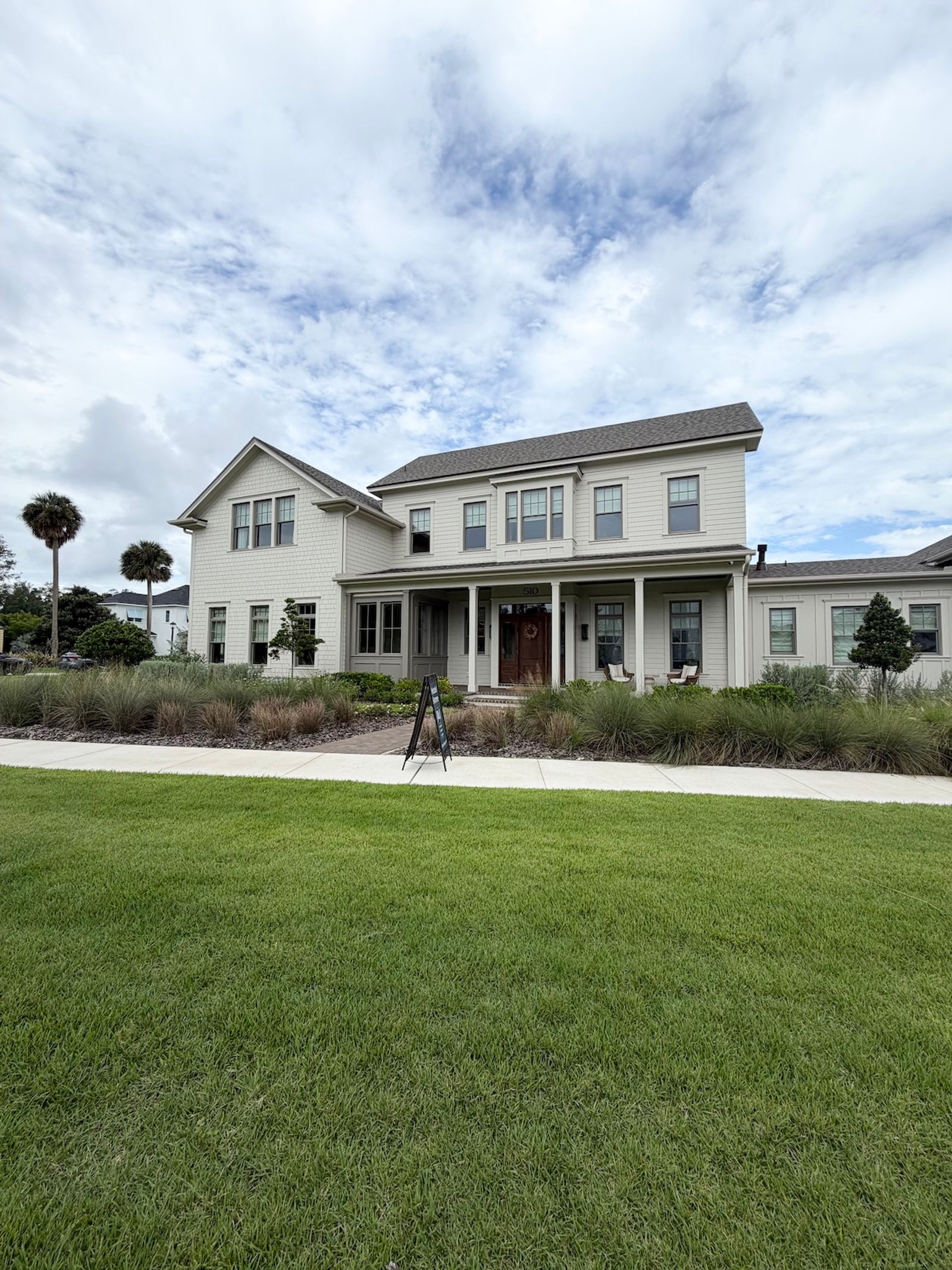 Two-story white farmhouse-style home with a covered front porch, dark wood front door, and landscaped yard with palm trees under a partly cloudy sky