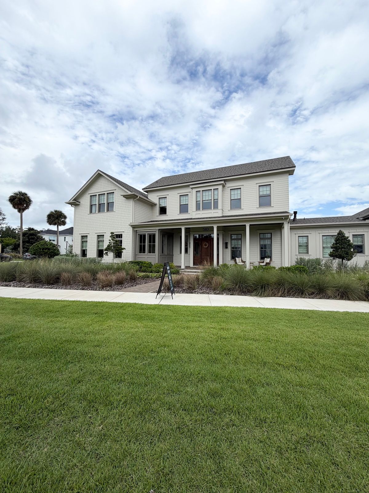 Two-story white farmhouse-style home with a covered front porch, dark wood front door, and landscaped yard with palm trees under a partly cloudy sky