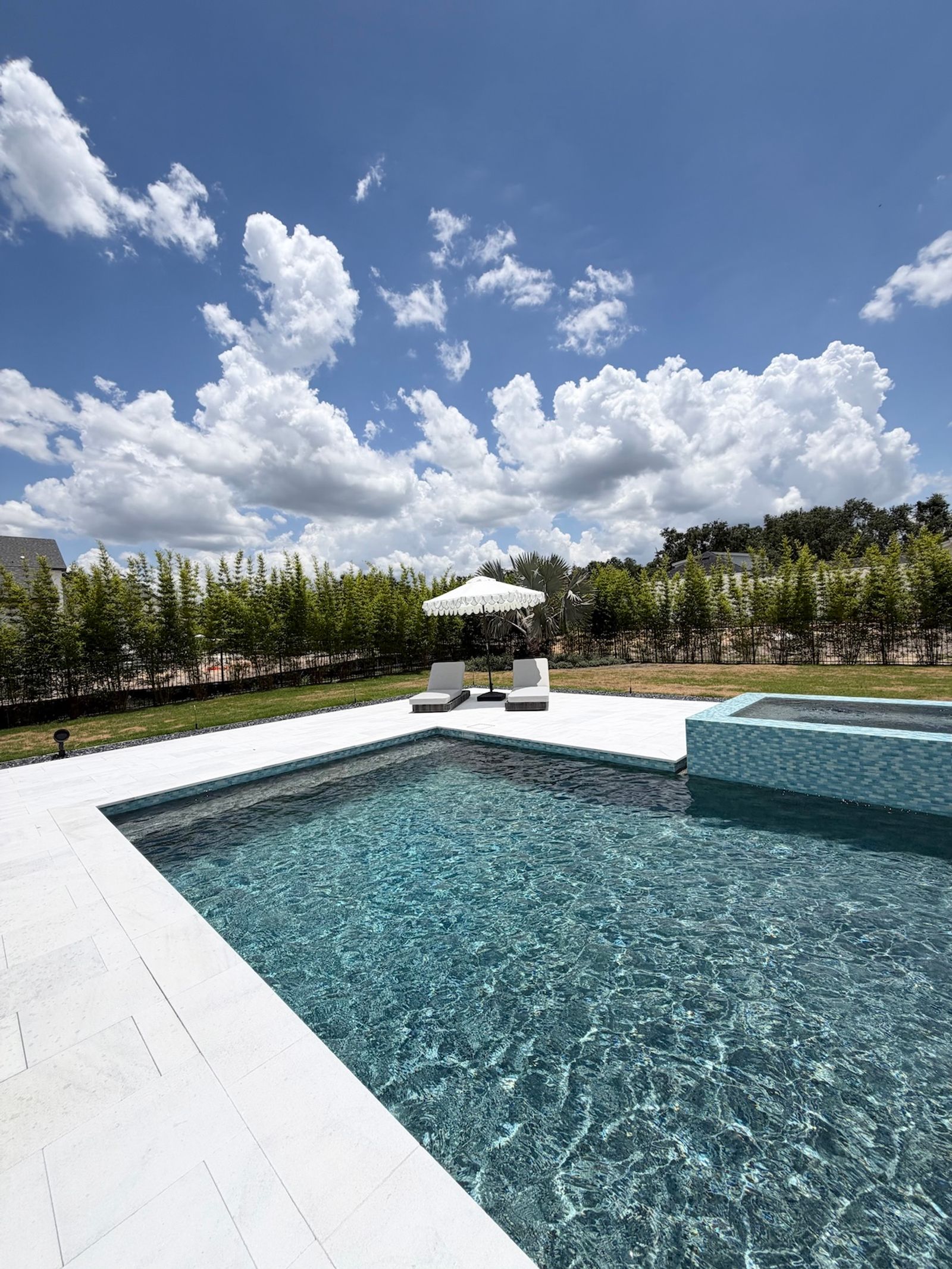 Backyard pool with blue-gray tile and a raised spa, surrounded by white pavers, with two lounge chairs under a white umbrella and a row of young trees along the lawn