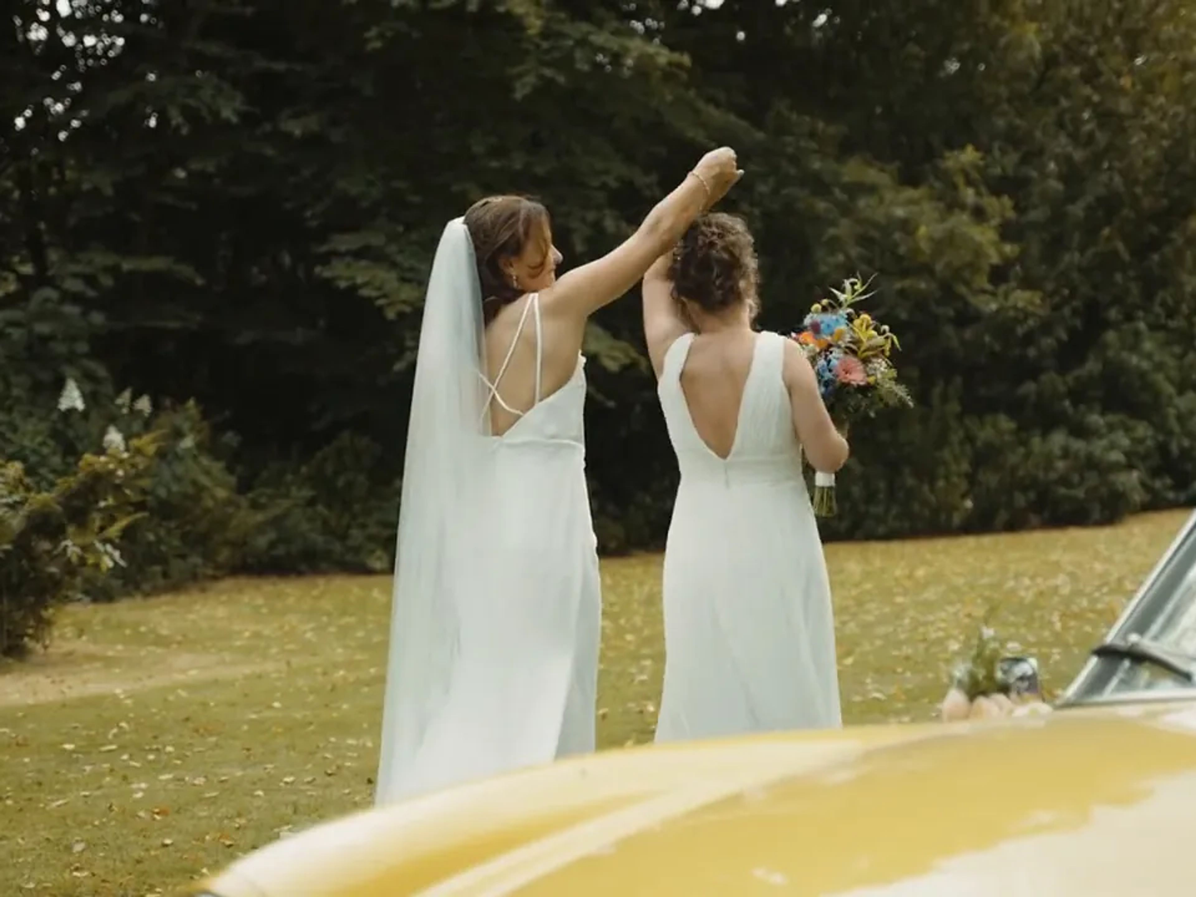 Two brides cheering with their bouquet next to a yellow convertible