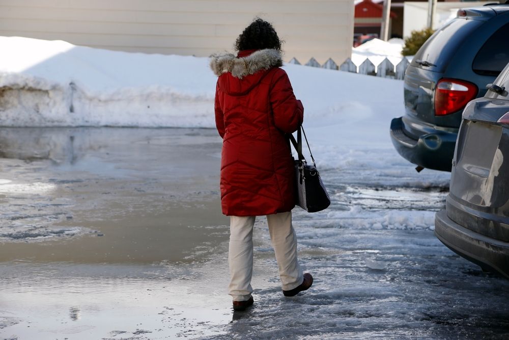 A person in a red winter parka walking across a slushy, icy parking lot between parked cars during the winter holiday shopping season.