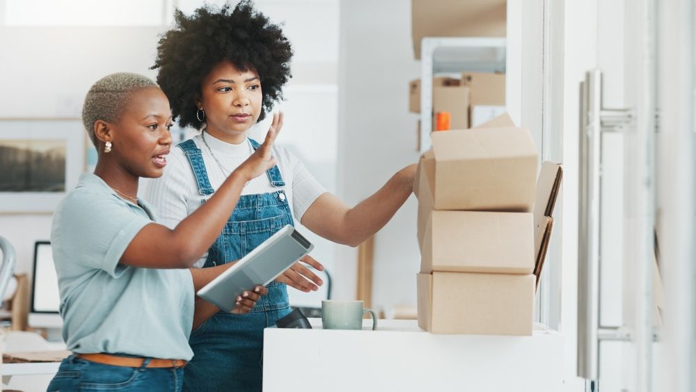 Two retail workers in a stockroom using a tablet to manage an overflowing stack of holiday shipment boxes.