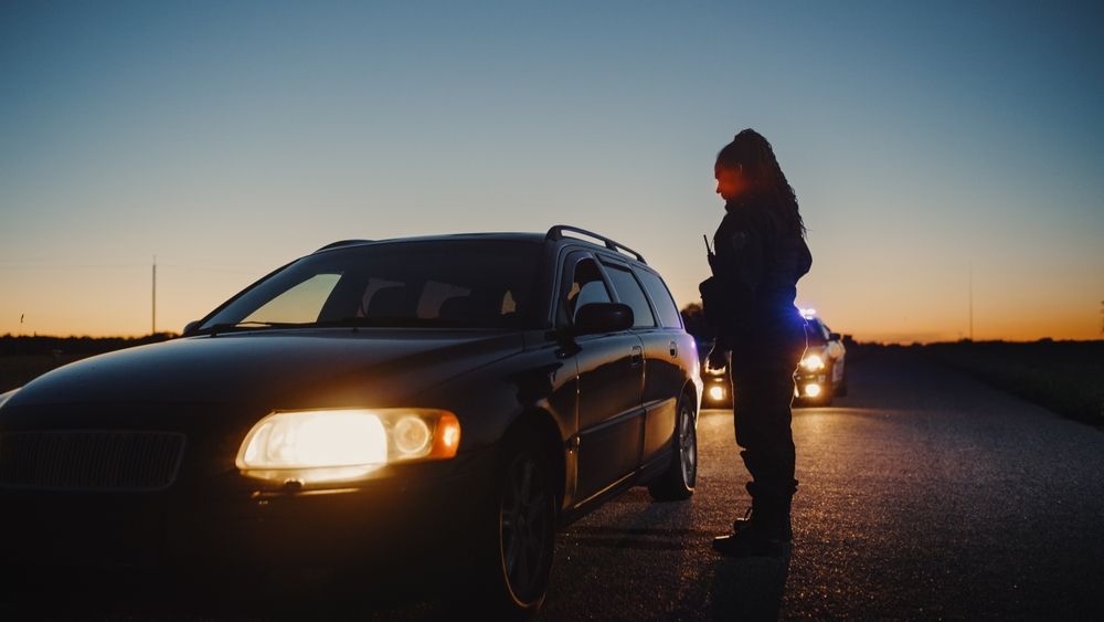 Police officer stands beside a pulled over car at dusk while patrol lights flash behind on a highway, symbolizing DUI stops following holiday parties.