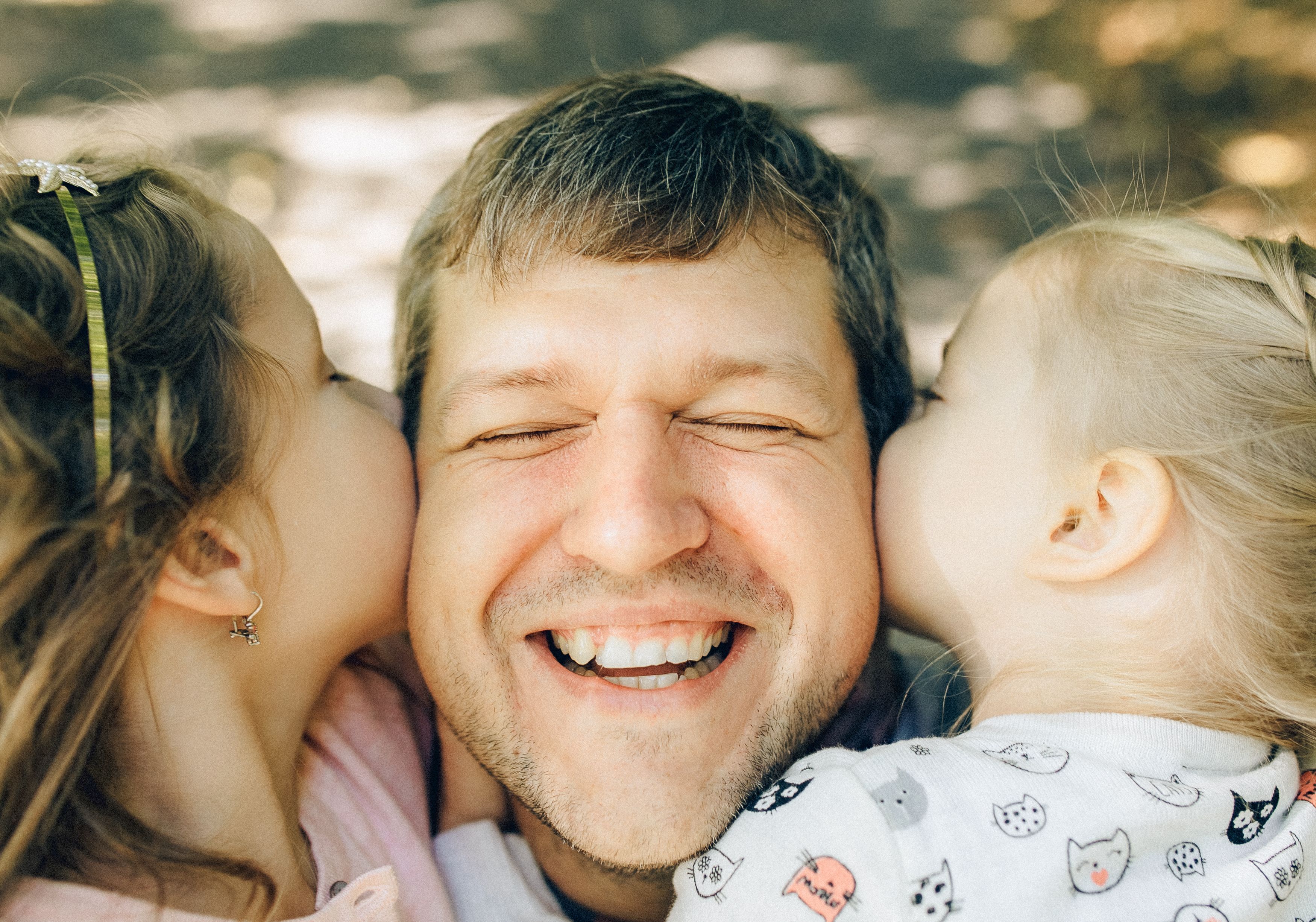 A laughing father is kissed on both cheeks simultaneously by his two young daughters outdoors, capturing a joyful Father's Day moment.