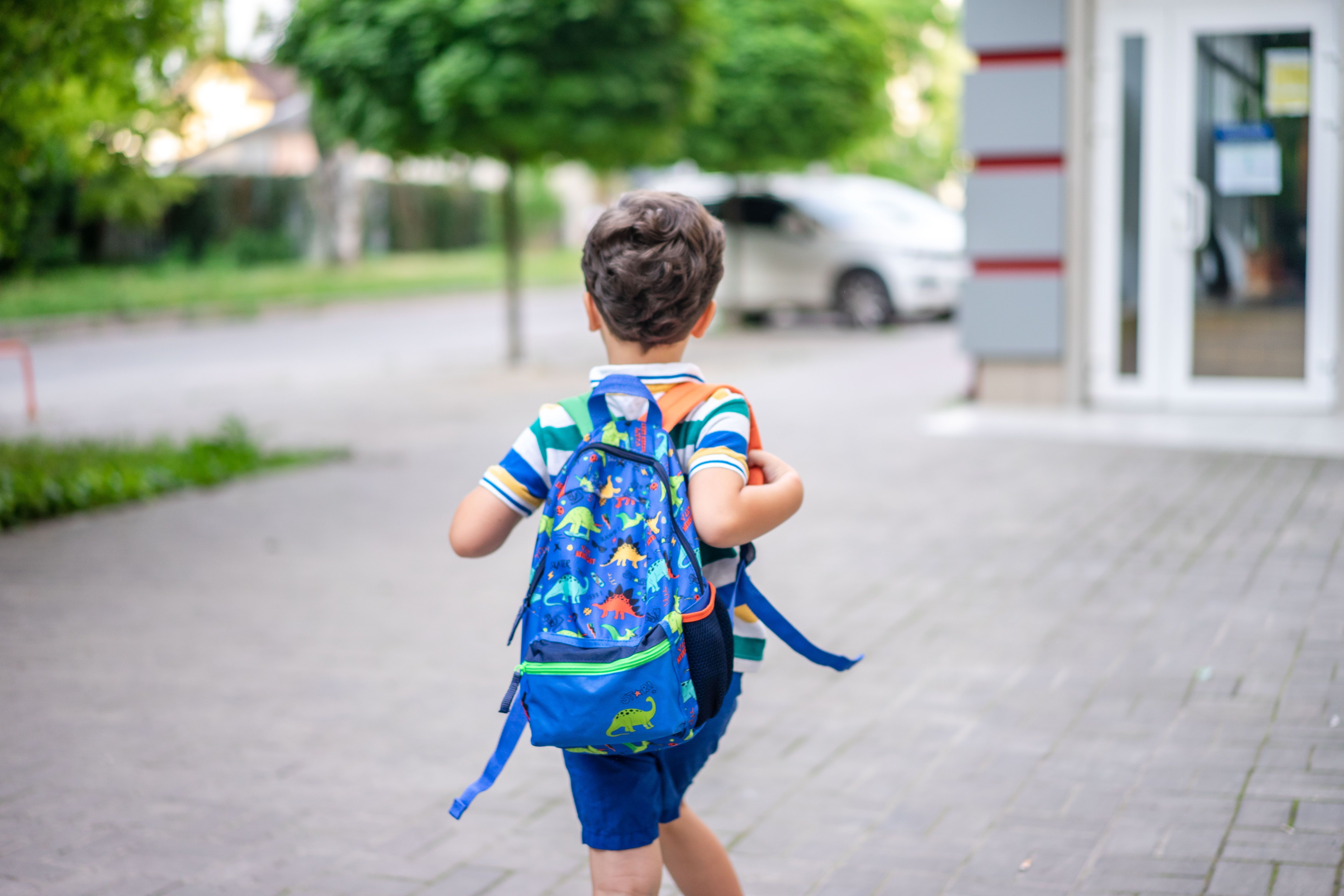 Young boy walking to school alone wearing a dinosaur backpack near a crosswalk, illustrating back-to-school pedestrian safety risks for New Jersey children.