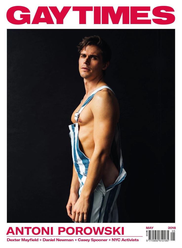 Antoni Porowski shirtless in striped denim overalls against a black backdrop on the cover of Gay Times, photographed by Taylor Miller.