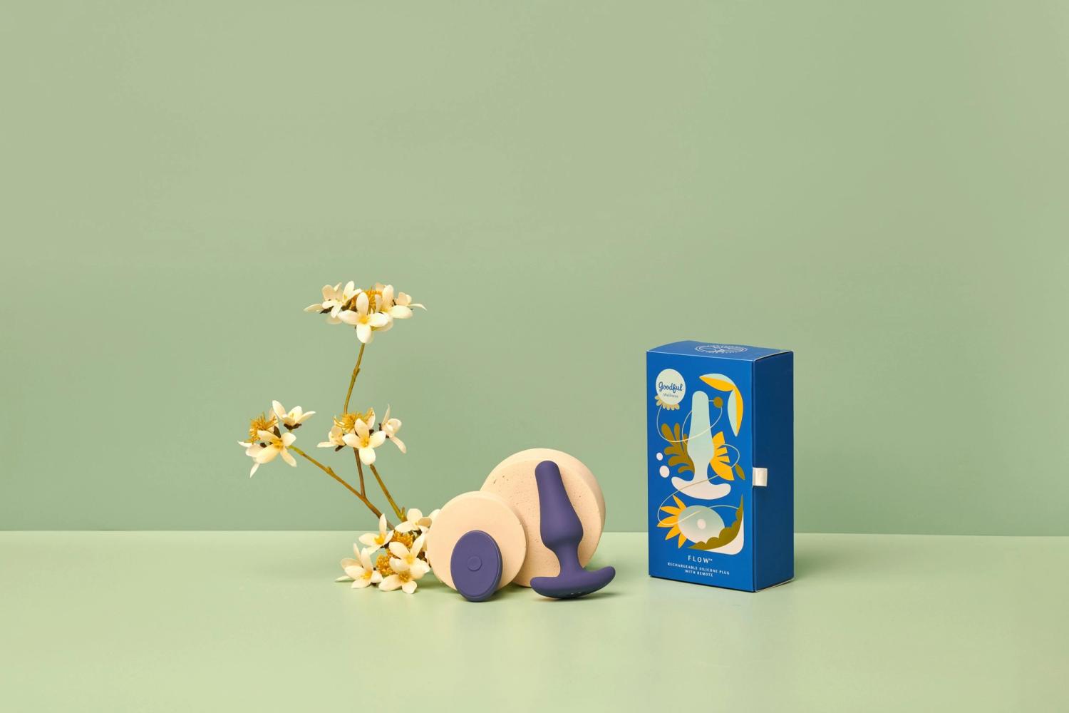 Goodful x Pipedream Flow silicone plug and remote beside its blue box and white blossoms on a green surface, photographed for BuzzFeed by Taylor Miller.