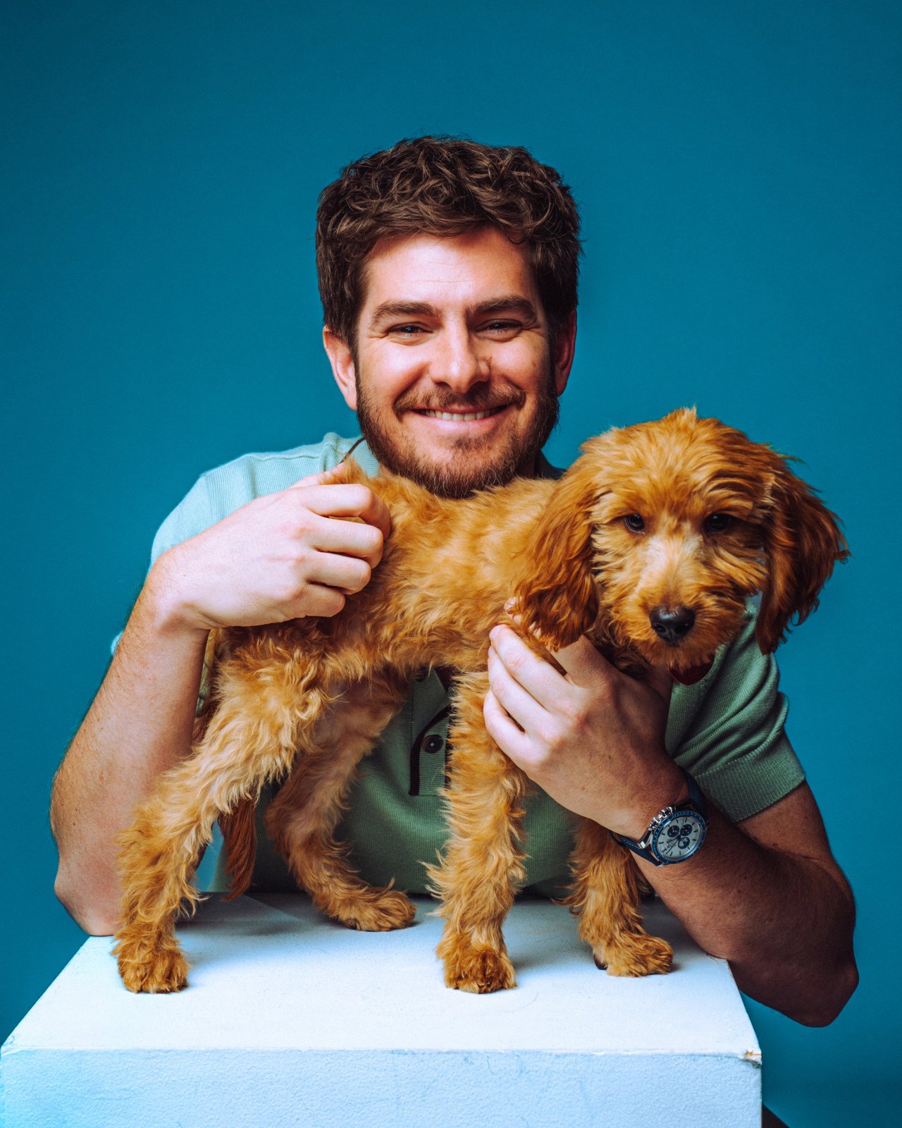 Andrew Garfield in a sage green polo smiling beside a fluffy apricot puppy against a teal backdrop, photographed by Taylor Miller.