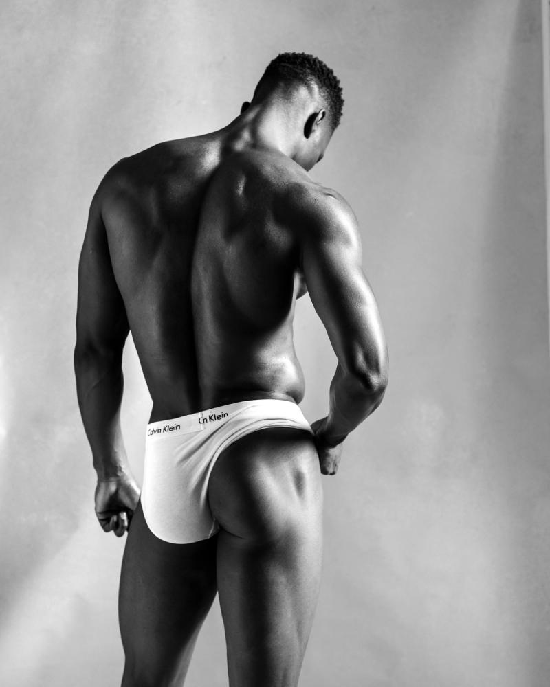 A muscular male model photographed from behind in white Calvin Klein briefs against a neutral studio wall, photographed by Taylor Miller.