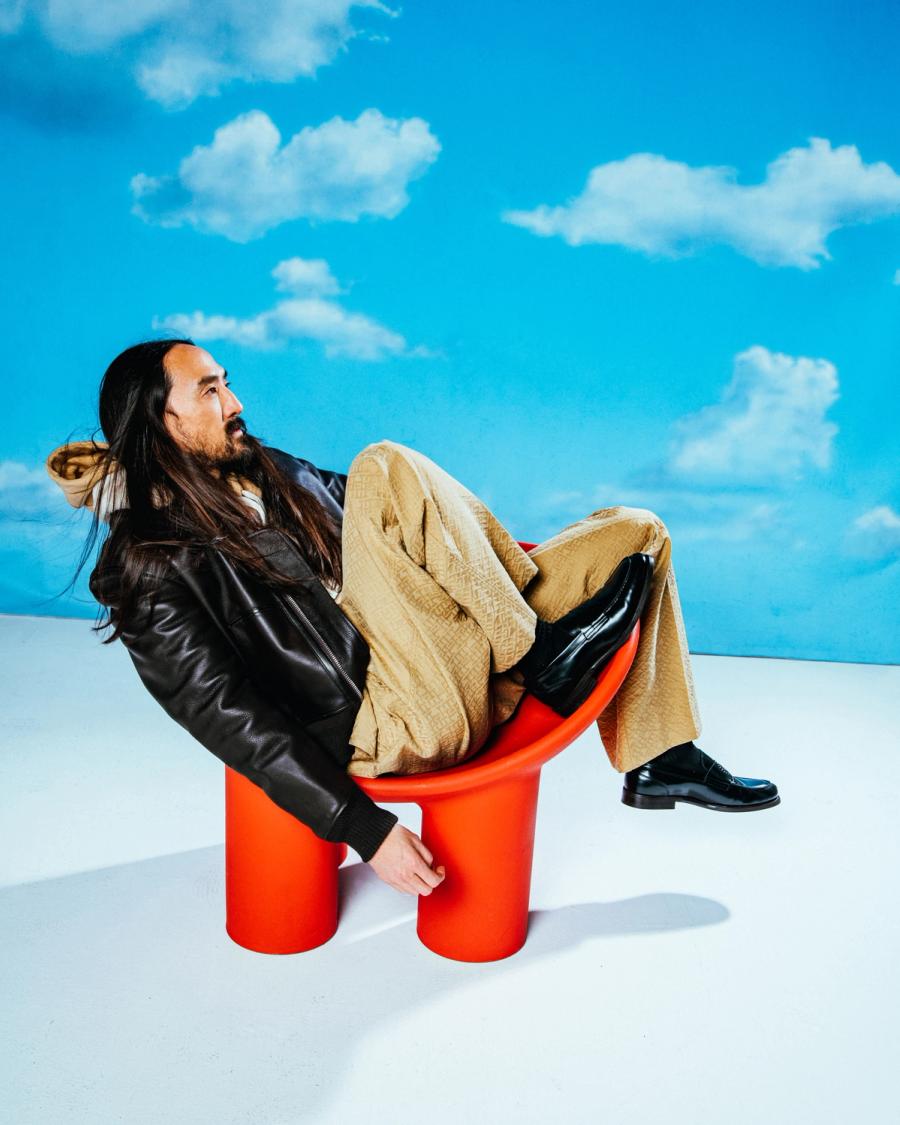 Steve Aoki, wearing a Tommy Hilfiger Jacket and Pants in front of a sky background on a red chair, photographed for Complex and Tommy Hilfiger by photographer Taylor Miller.
