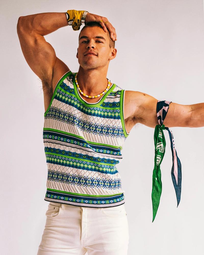 Mitchell Slaggert posing with one arm raised in a green and blue patterned knit tank and white trousers, photographed for Numéro Netherlands by Taylor Miller.