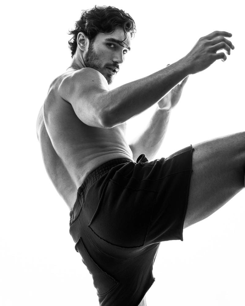Black and white frame of Michael Yerger, shirtless in dark shorts with one leg kicked high, for L'Officiel Hommes by photographer Taylor Miller.