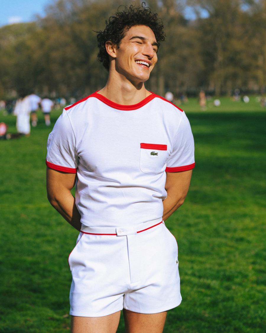 Sacha Bilal in a white Lacoste tee with red trim and matching shorts in a sunny park, Lacoste NYC 2026 campaign by Taylor Miller.