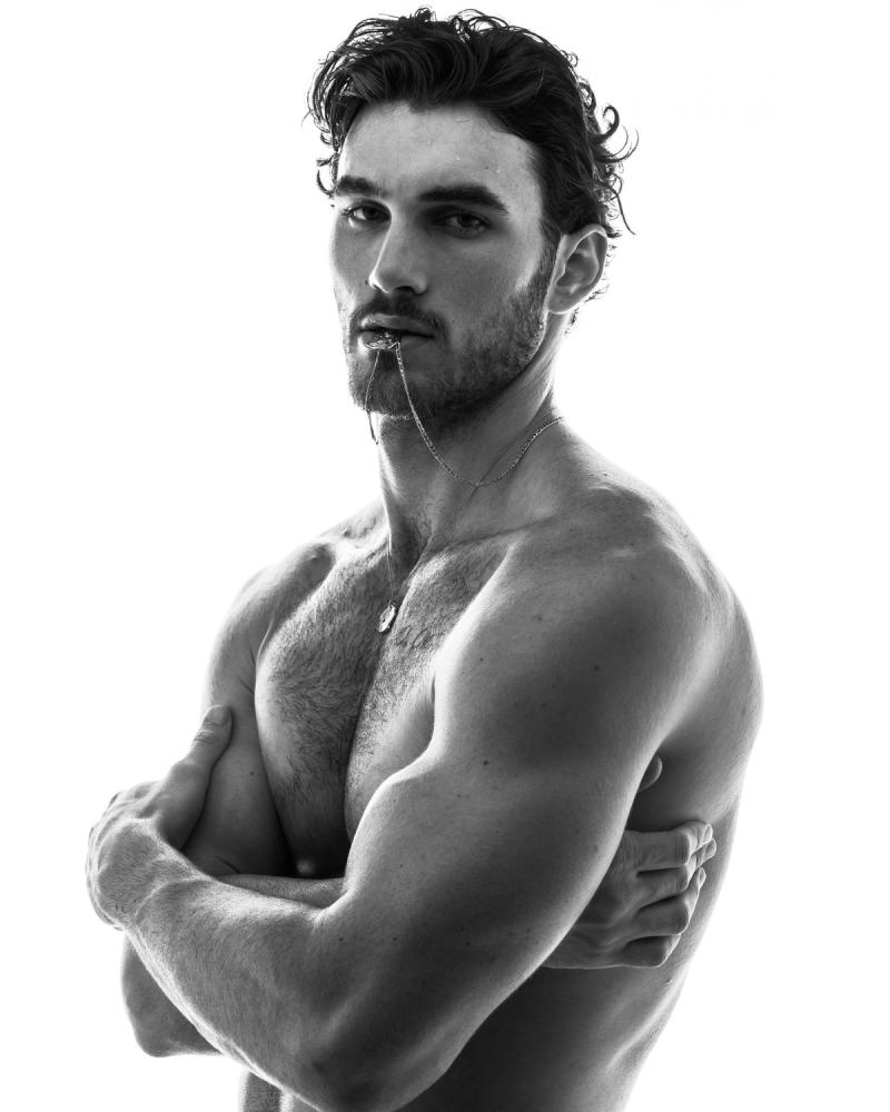 Black and white portrait of Michael Yerger, arms crossed and a silver chain held in his teeth, for L'Officiel Hommes by photographer Taylor Miller.