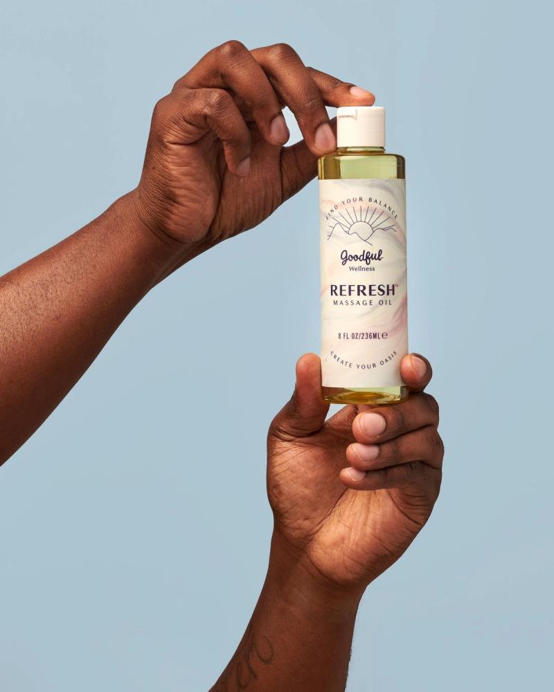Hands holding a bottle of Goodful Wellness Refresh Massage Oil against a blue background, photographed for BuzzFeed by Taylor Miller.