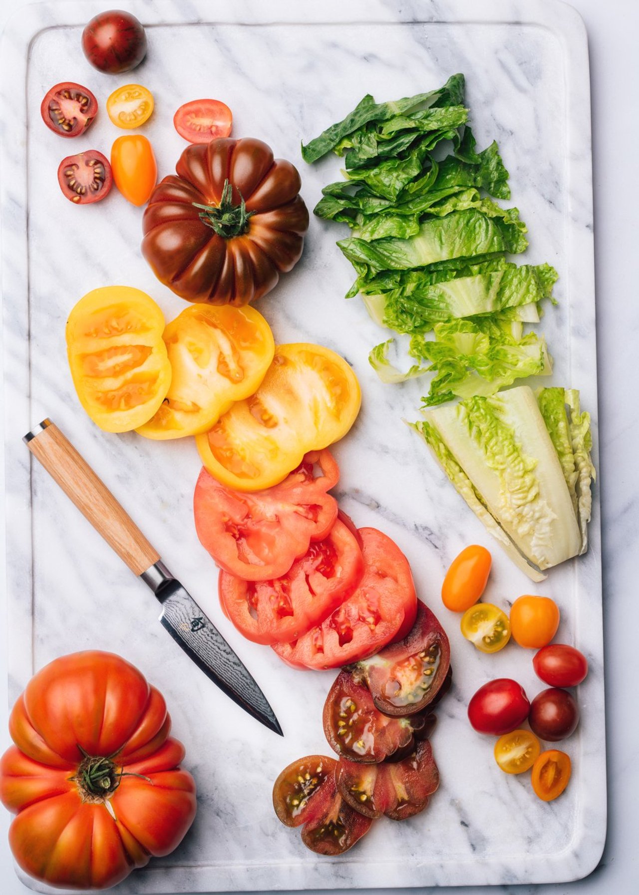 Kai damascus knife on a marble board with sliced red and yellow heirloom tomatoes and chopped romaine lettuce, photographed for BuzzFeed by Taylor Miller.