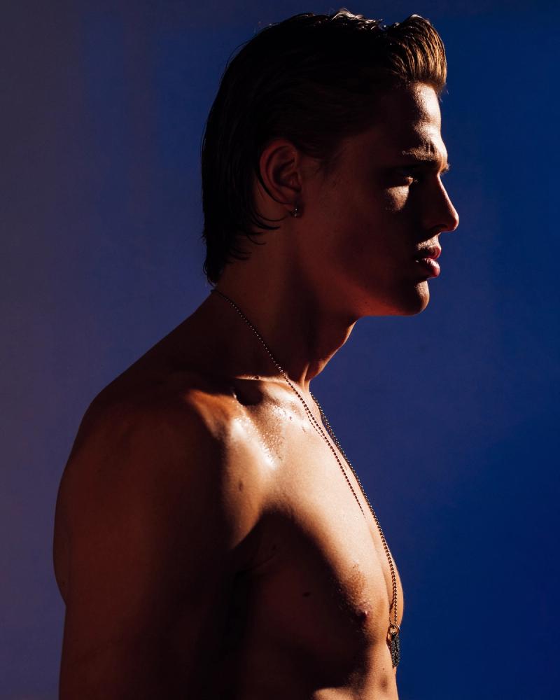 Taras Romanov in profile with slicked-back hair and a long chain necklace against a blue backdrop, photographed by Taylor Miller.