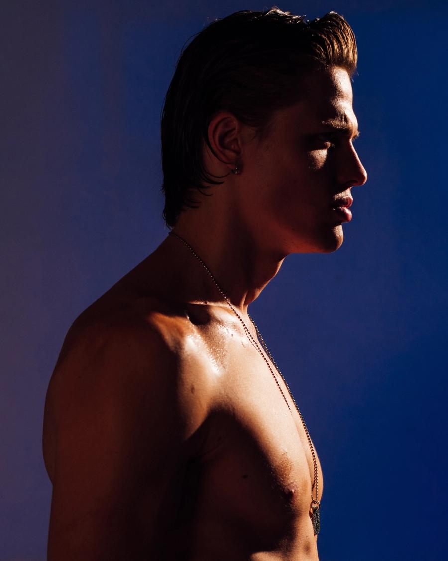 Taras Romanov in profile with slicked-back hair and a long chain necklace against a blue backdrop, photographed by Taylor Miller.