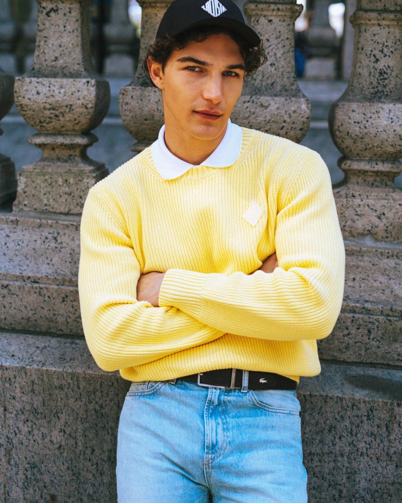 Sacha Bilal in a yellow ribbed Lacoste sweater, jeans and black cap leaning on stone, for the Lacoste NYC 2026 campaign by Taylor Miller.
