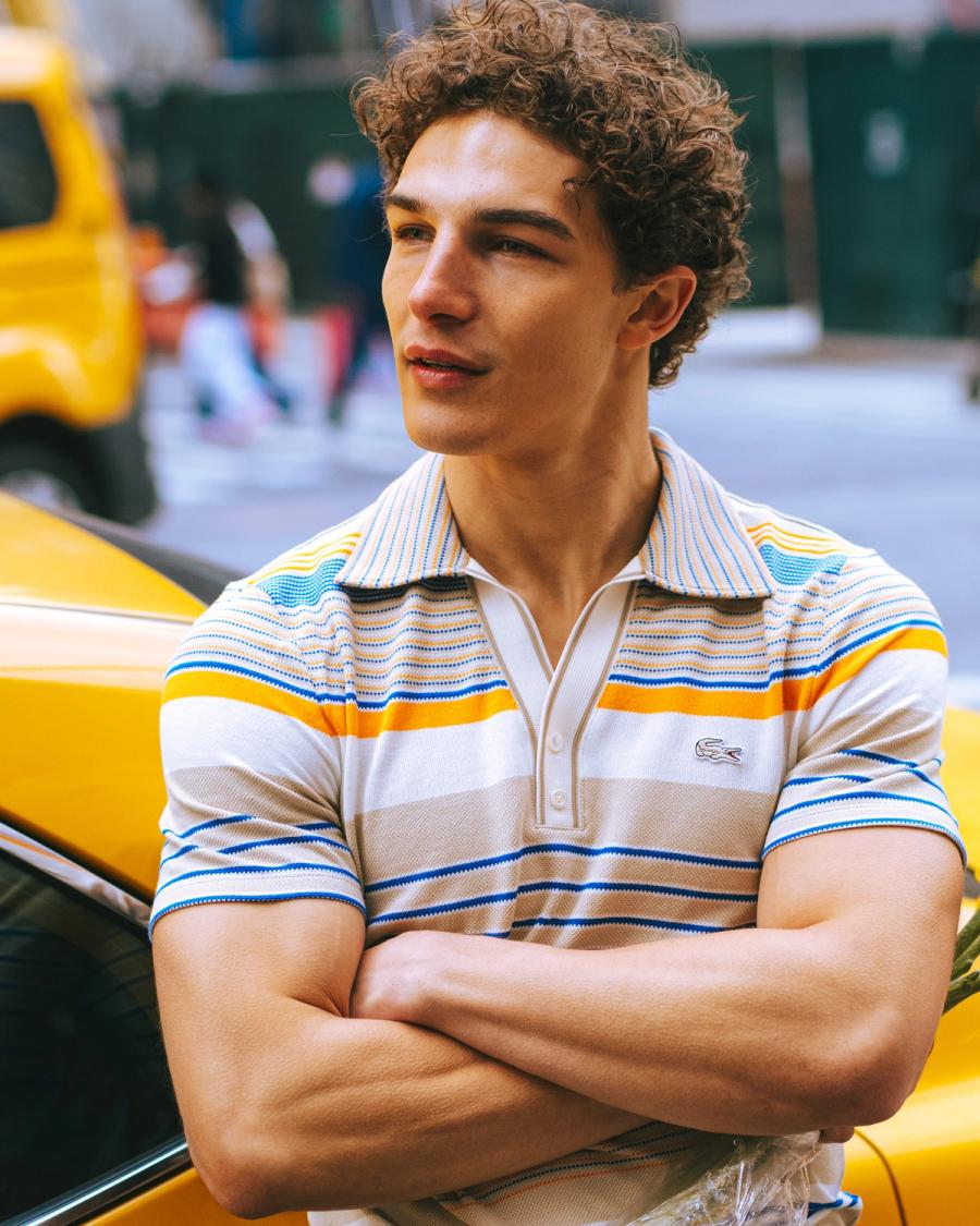 Sacha Bilal in a striped Lacoste polo leans against a yellow taxi with arms crossed, for the Lacoste NYC 2026 campaign by photographer Taylor Miller.