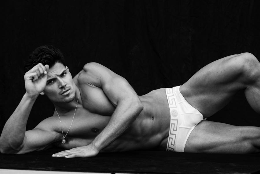 William Goodge reclining in white Versace briefs against a black backdrop in a black and white portrait, photographed by Taylor Miller.