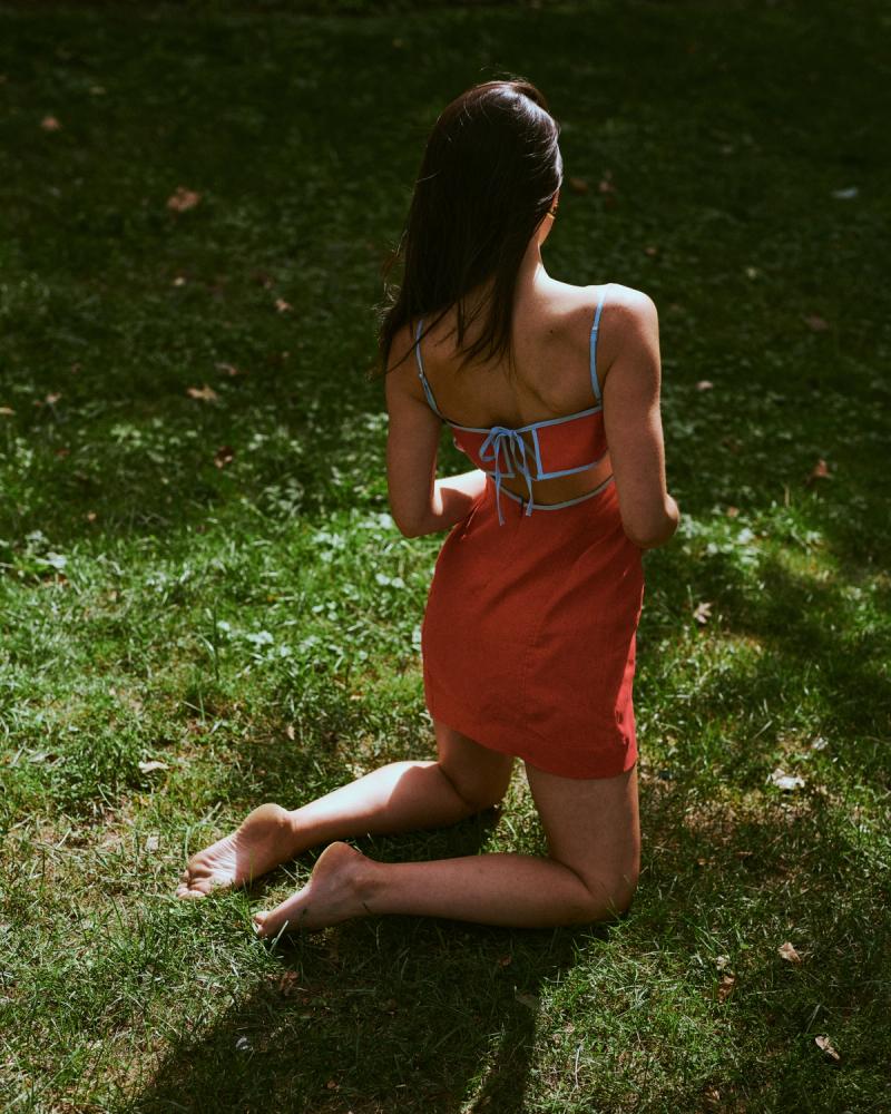 Rear view of Mary Holland Nader kneeling on grass in a red linen dress with blue tie-back detail, ESYA campaign photographed by Taylor Miller.