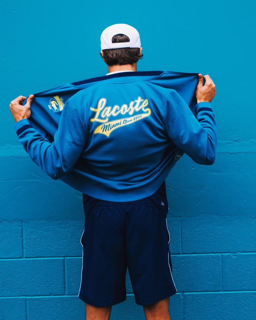 From behind, a man opens a blue jacket revealing the yellow Lacoste Miami Open 2025 script, photographed by Taylor Miller.