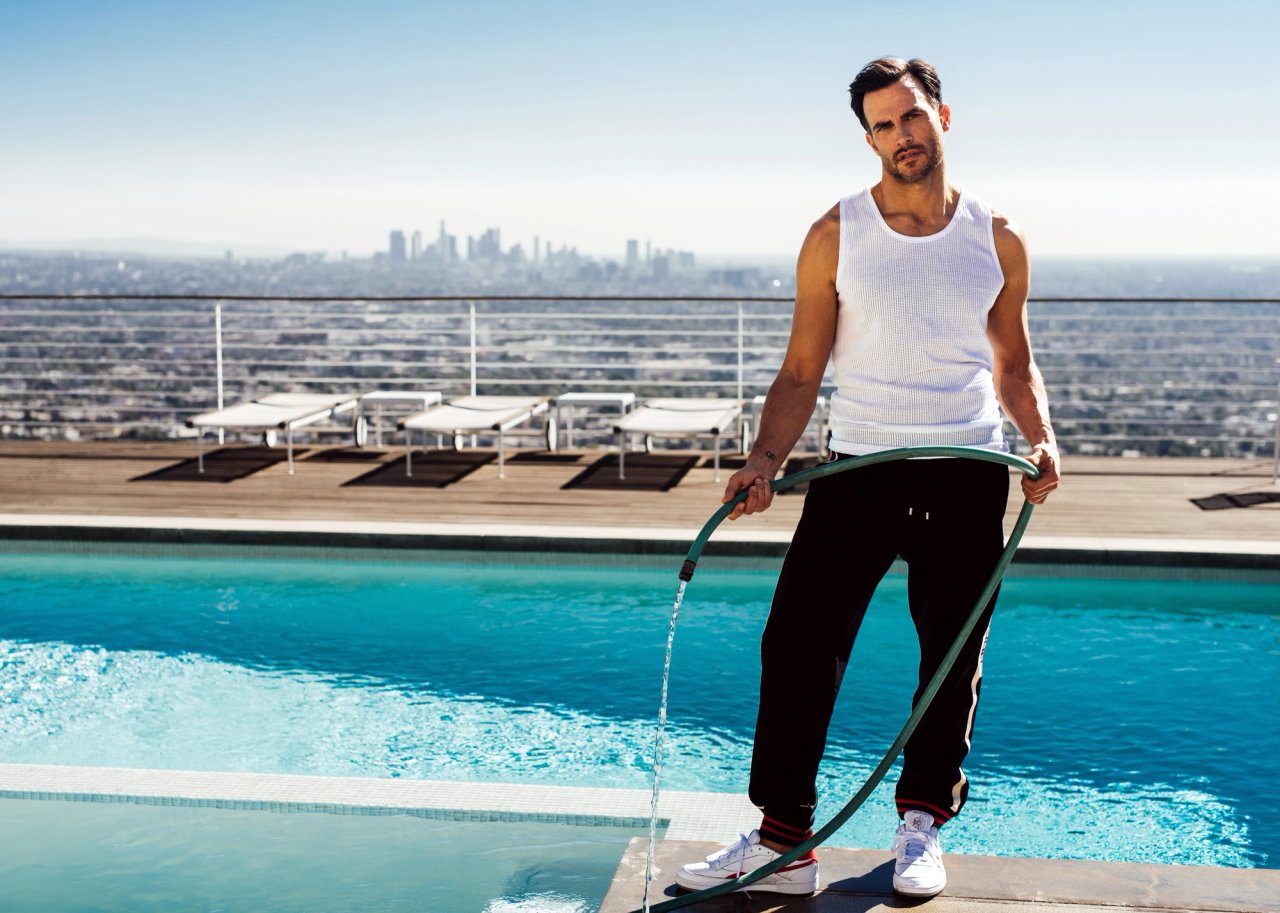 Cheyenne Jackson in a white tank and black track pants holding a running hose by a rooftop pool overlooking the Los Angeles skyline, photographed by Taylor Miller.
