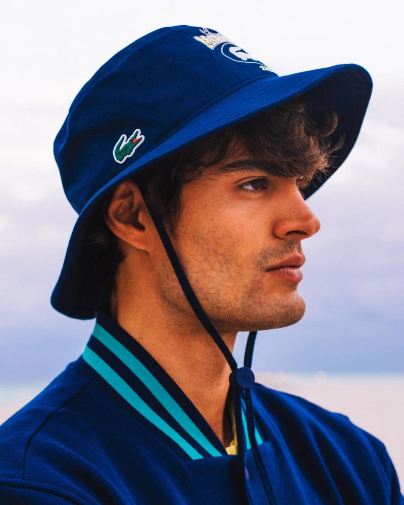 Profile of Iago Botelho in a navy Lacoste bucket hat and striped polo by the beach, for Lacoste Miami Open 2025 by Taylor Miller.