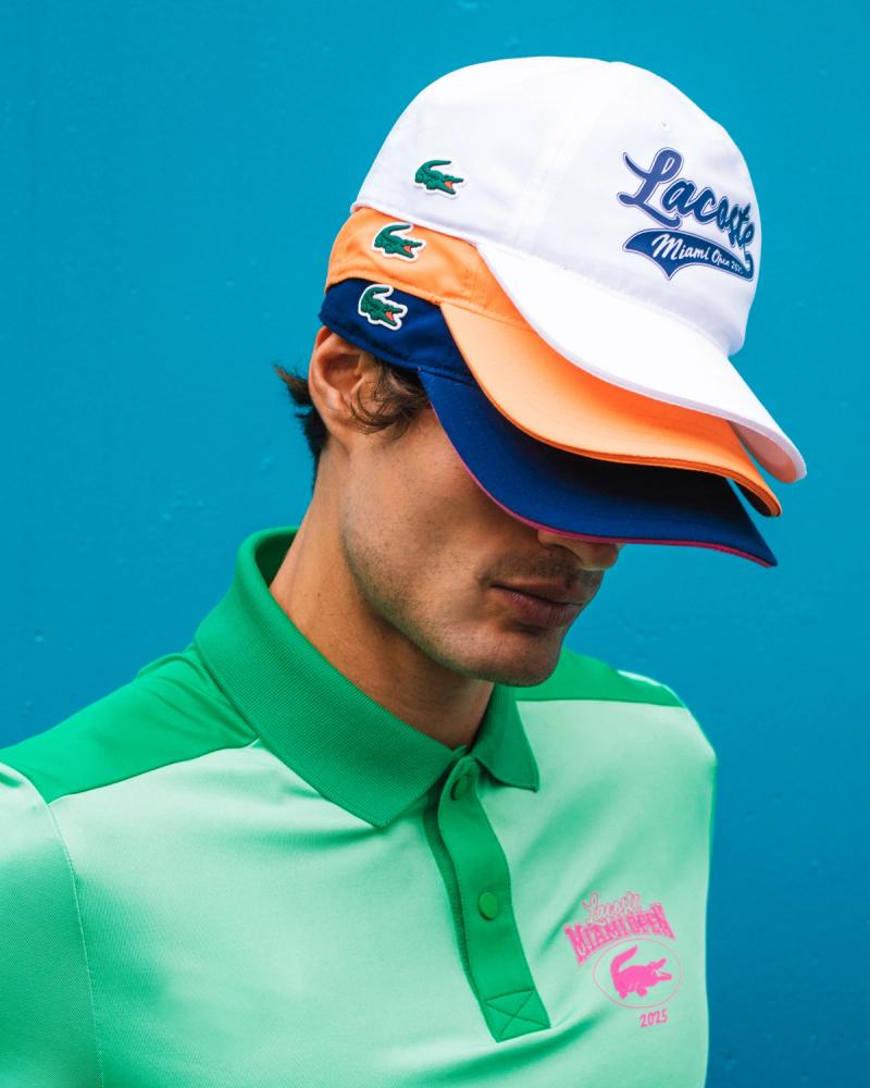 Iago Botelho in a green Lacoste polo wearing three stacked Lacoste Miami Open 2025 caps against a blue wall, by Taylor Miller.