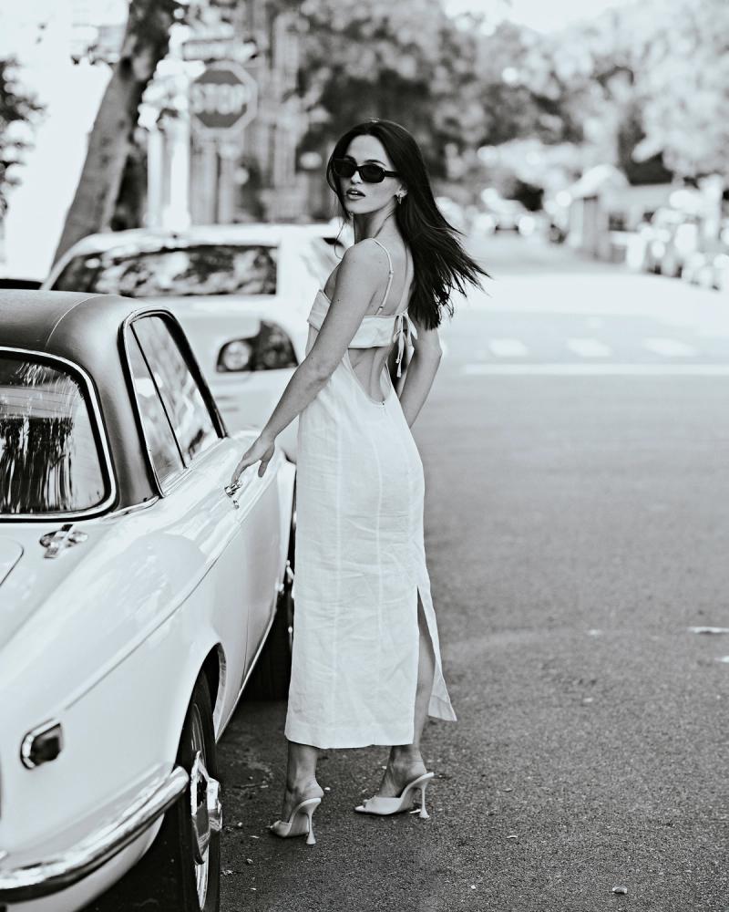 Black-and-white shot of Mary Holland Nader in a white midi slip dress and sunglasses beside a classic car for the ESYA campaign by Taylor Miller.