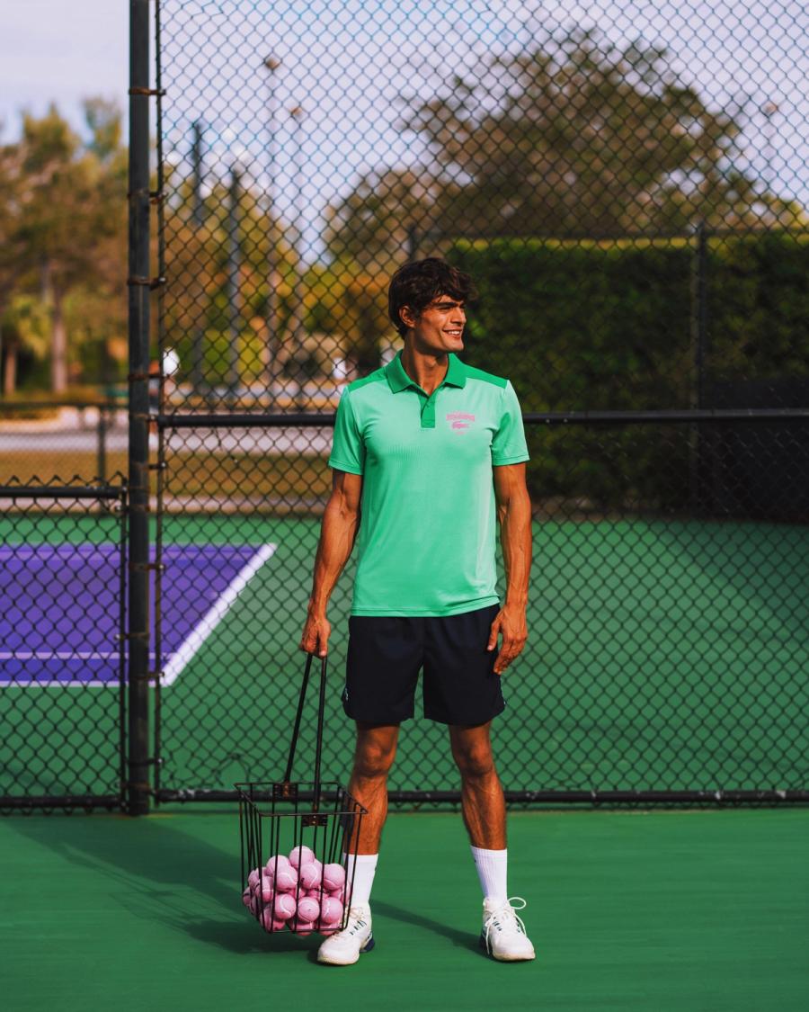 Iago Botelho in a mint green Lacoste polo holds a hopper of pink tennis balls courtside, for Lacoste Miami Open 2025 by Taylor Miller.